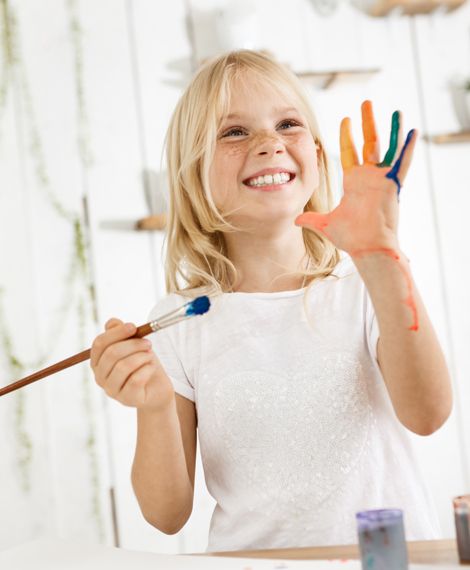 A smiling child with paint on her hand, holding paintbrushes